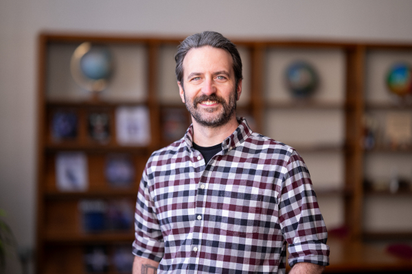 Photo of Nick Freed in a library smiling in a red/black/white plaid shirt