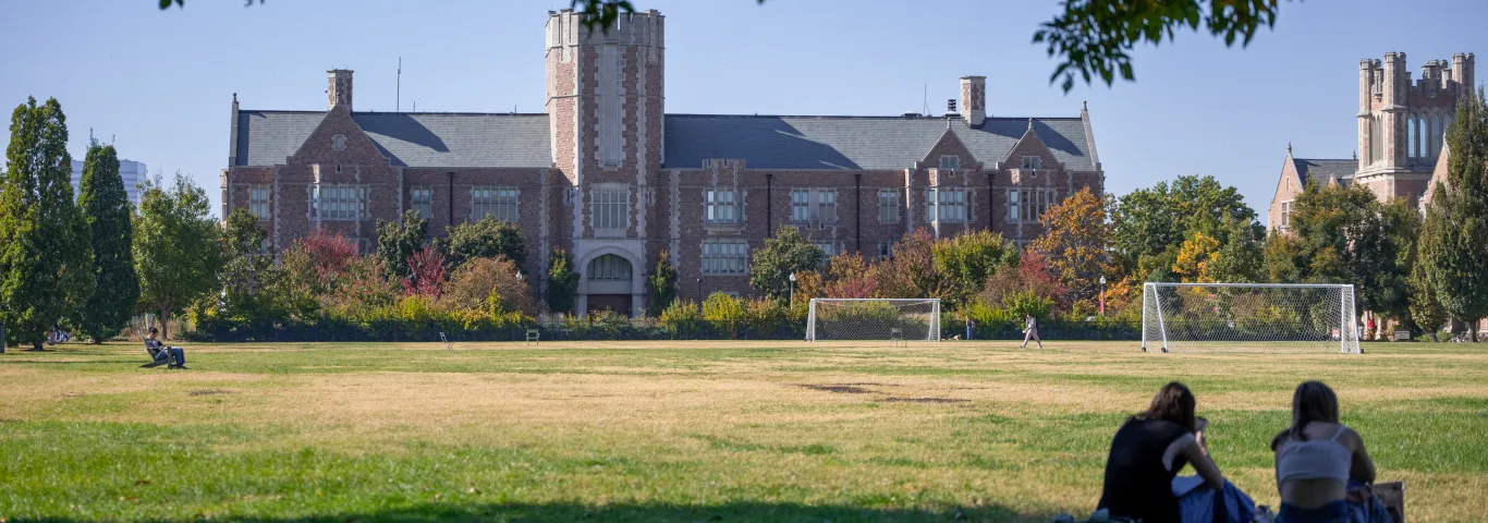 Photo of Seigle Hall during the day with two students in the bottom right corner sitting on the grass