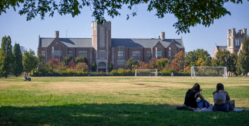 Photo of Seigle Hall during the day with two students in the bottom right corner sitting on the grass