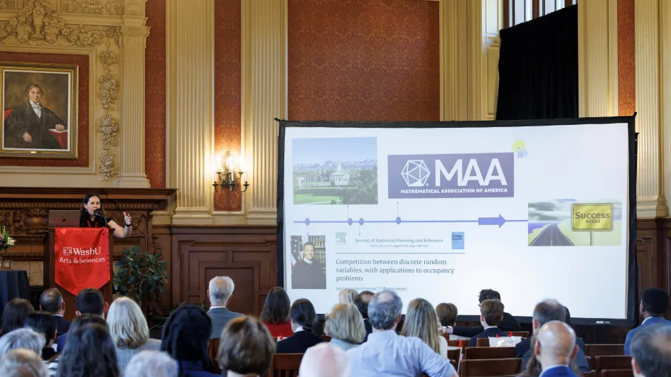 Photo of Betsy Sinclair giving a speech in front of an audience with a projector screen to her left displaying a slide