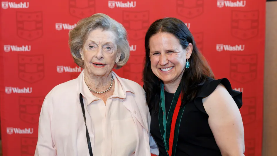 Photo of Betsy Sinclair and Barbara Ann Smith smiling to camera in front of a red WashU background