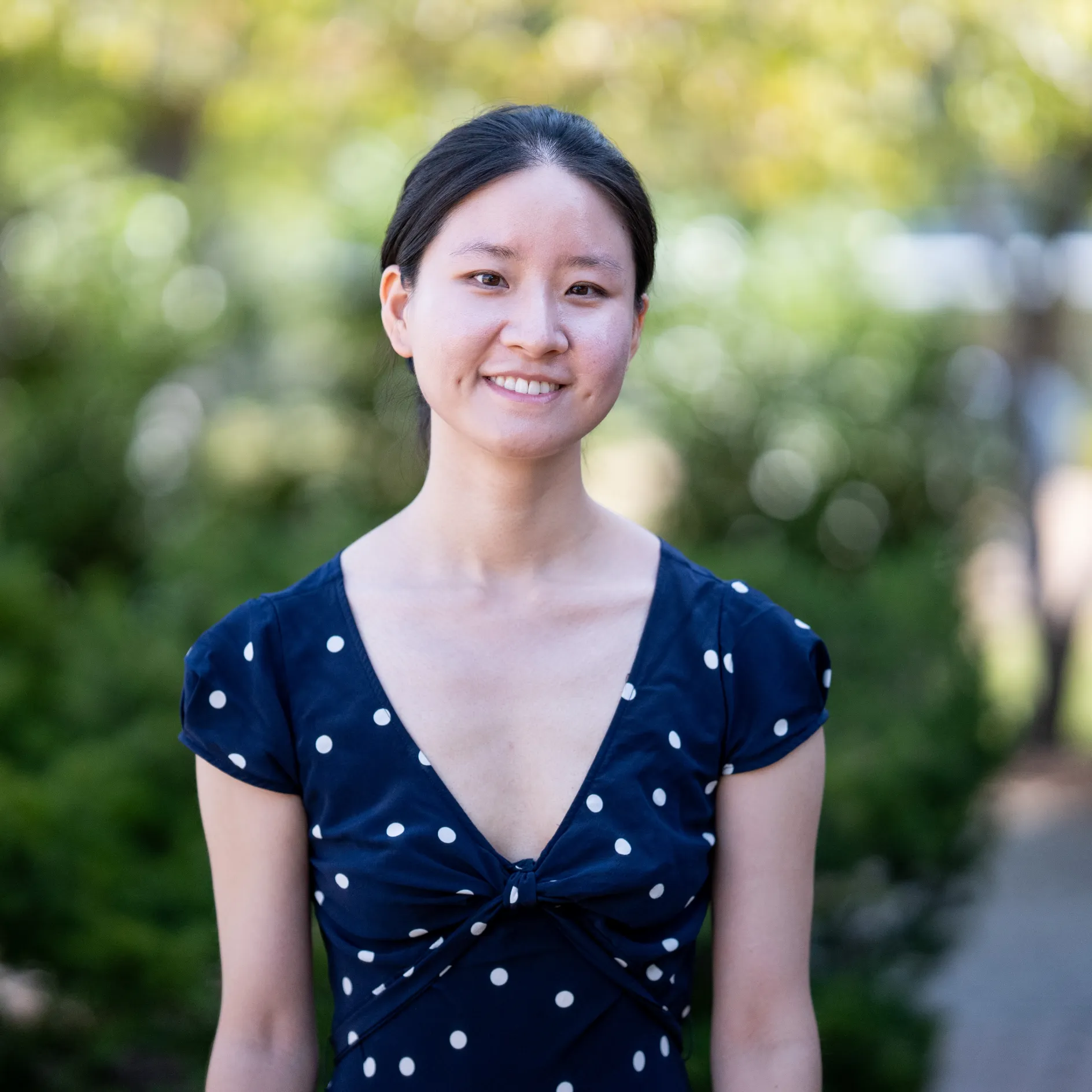 Photo of Jie Ma smiling to camera. They have dark hair in a pony tail and are wearing a blue top with white spots.