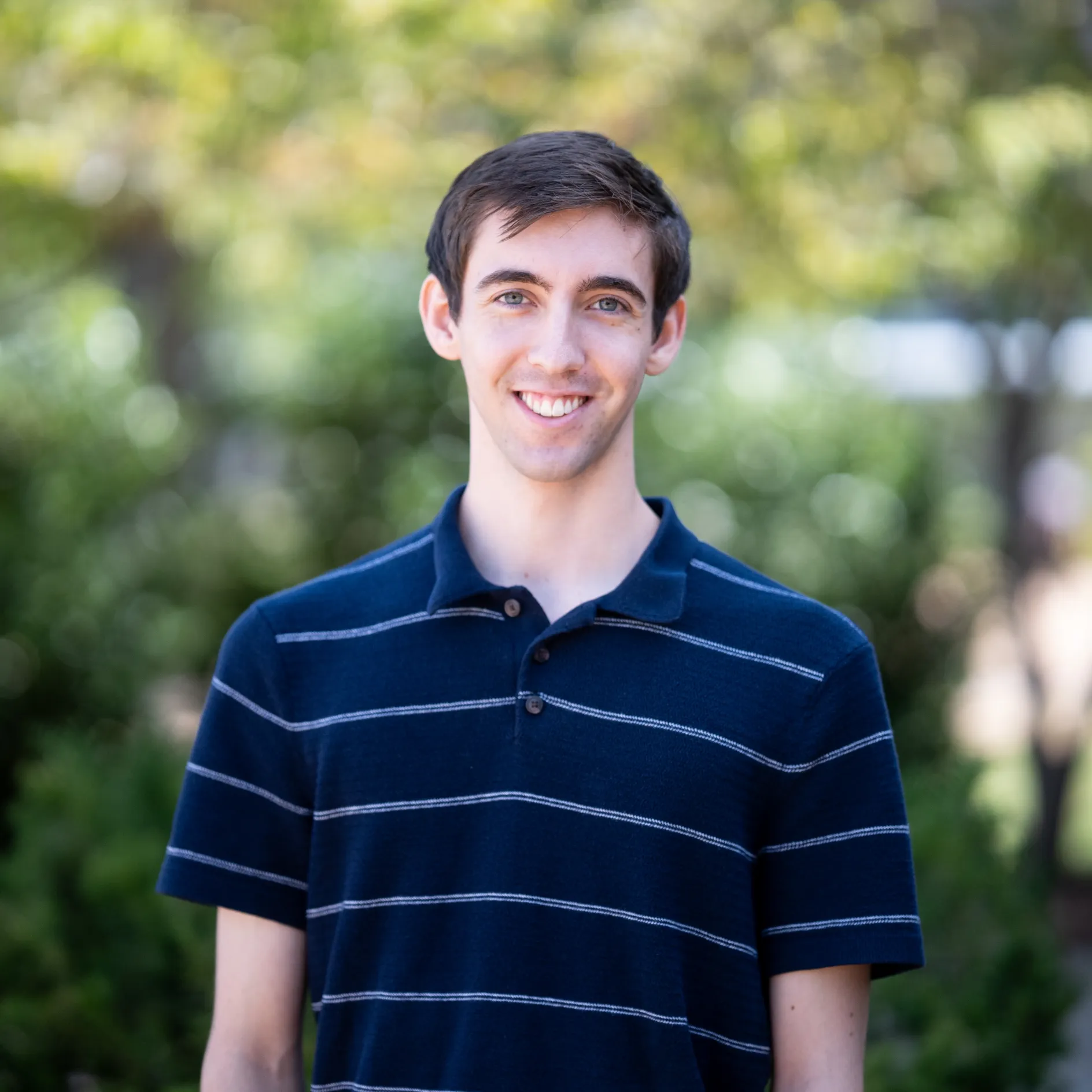 Photo of Ryan Rohrbach smiling to camera. They have short dark hair and are wearing a striped short sleeve polo shirt