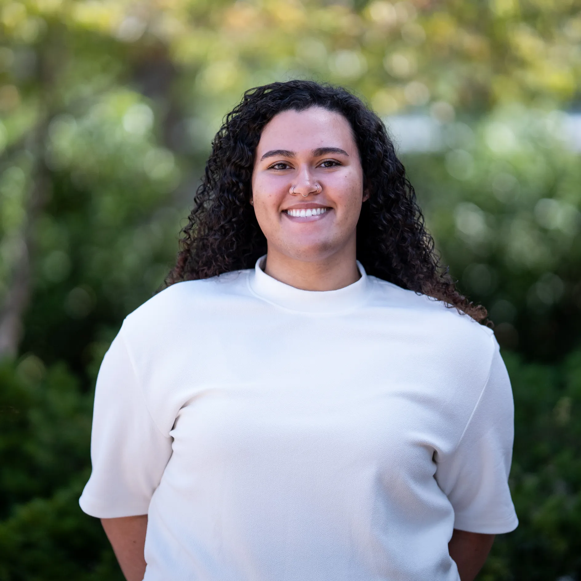 Photo of Jayden Sheridan smiling to camera. She has long, curly black hair and is wearing a white short sleeve shirt