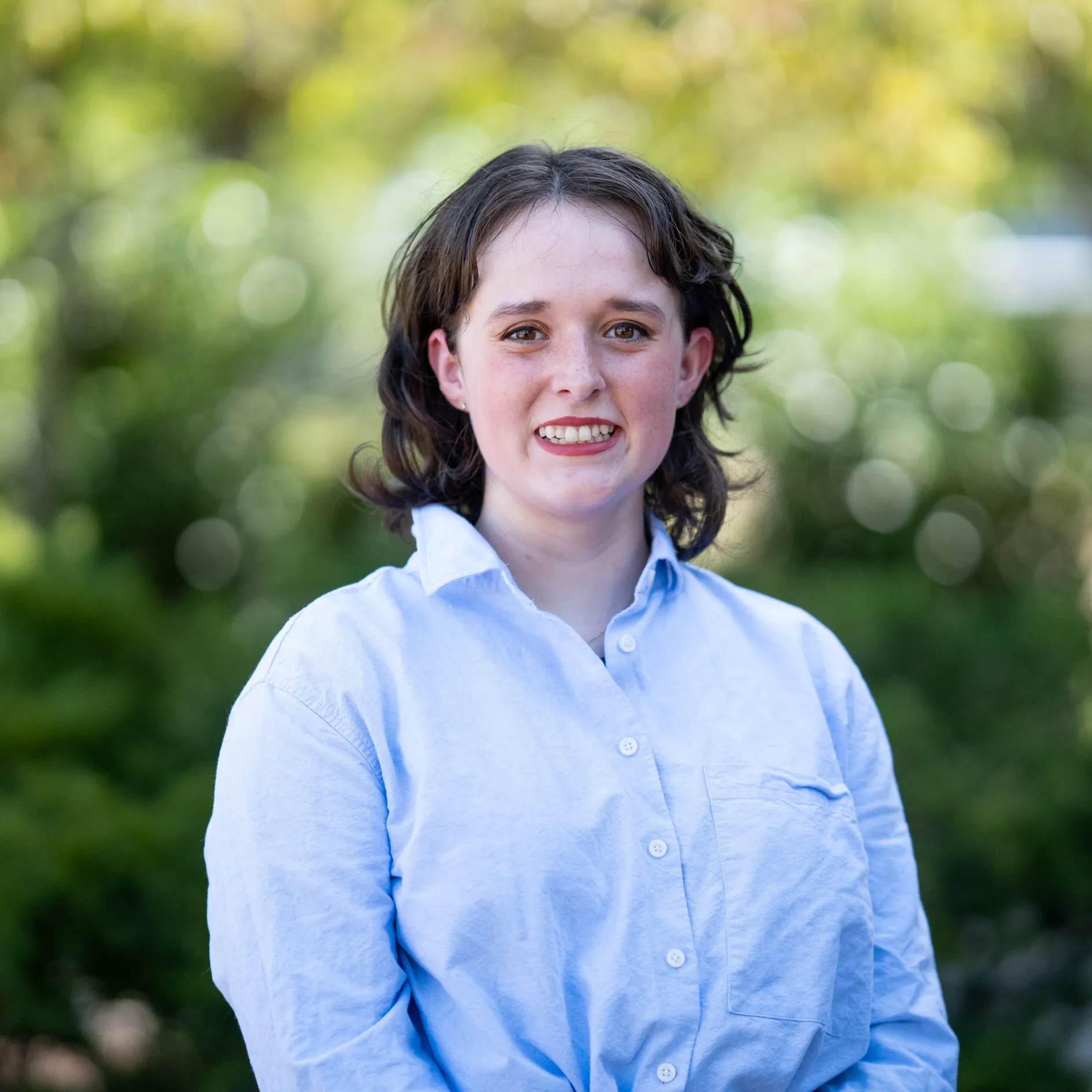 Photo of Addison "Rose" Wellenberger smiling to camera. They have short dark hair and are wearing a light blue button-up shirt