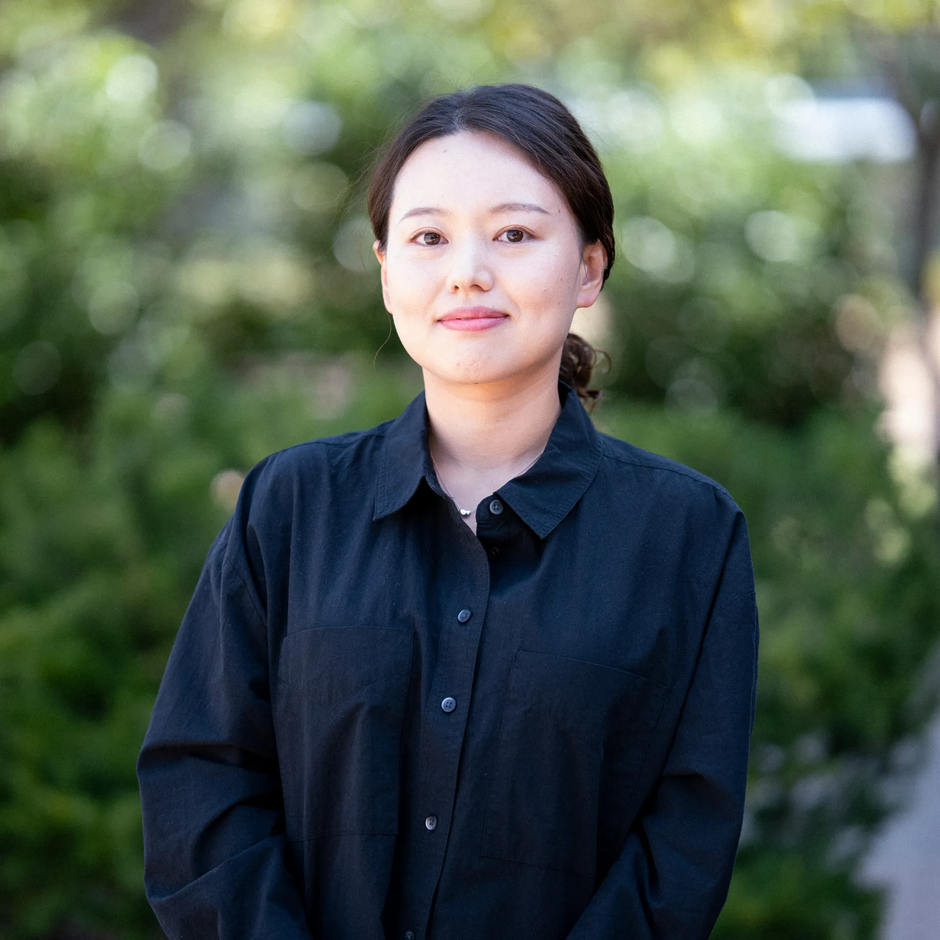 Photo of Seonkyu Pyo smiling to camera. They have long dark hair pulled back in a ponytail and are wearing a black button up shirt
