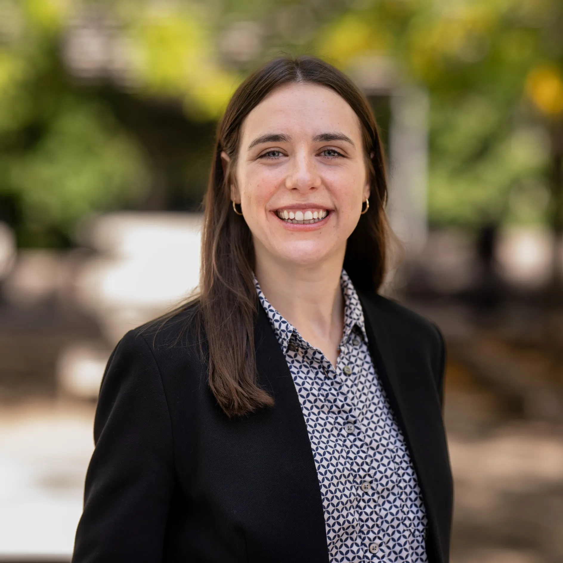 Photo of Ophelia Vedder, a white female-presenting person with shoulder length brown hair, smiling in a black blazer and gray top