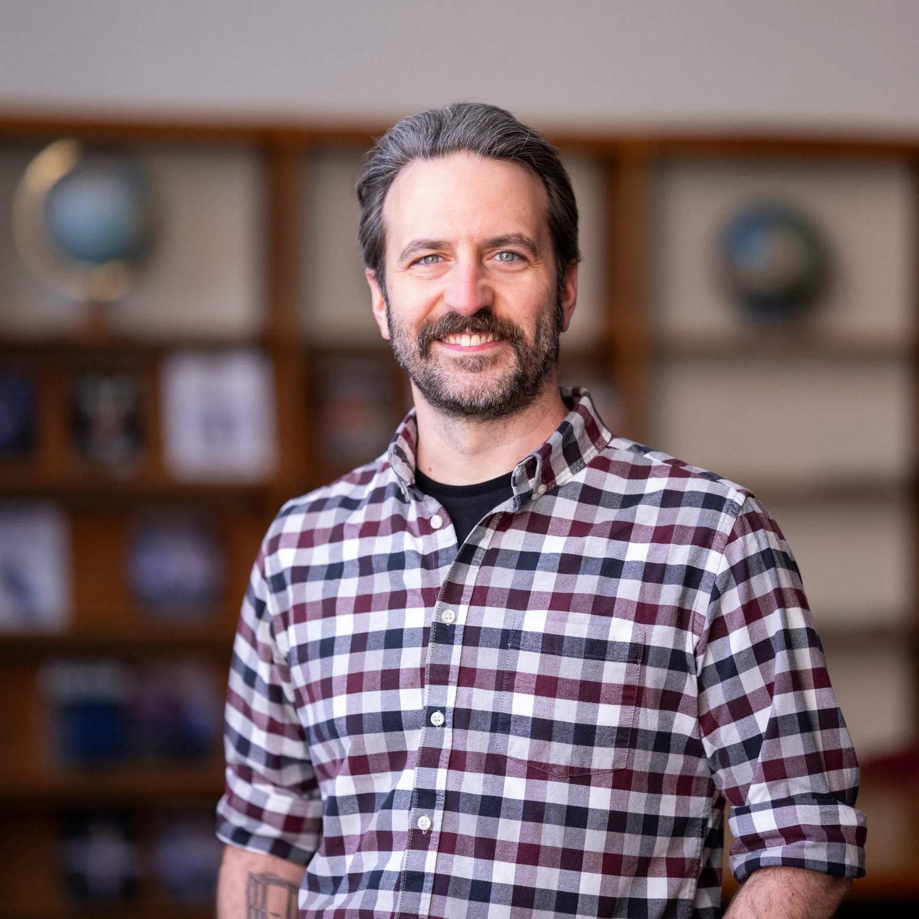 Photo of Nick Freed in a library smiling in a red/black/white plaid shirt