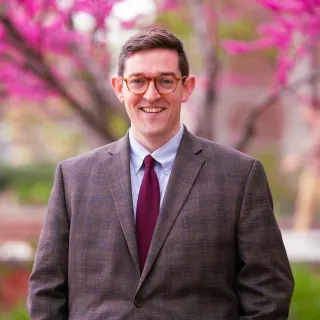 Photo of Michael Olson smiling to camera in a brown blazer, red tie, and blue button-up shirt