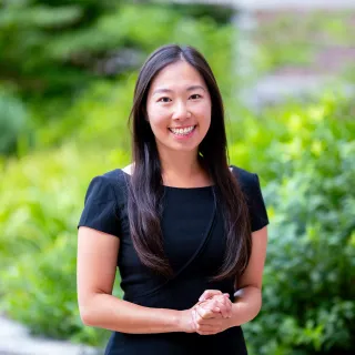 Photo of Elaine Yao smiling to the camera in front of greenery with hands clasped in front