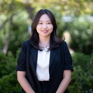 Photo of Jiin Ha smiling to camera. She has long black hair and is wearing a black jacket over a white top.