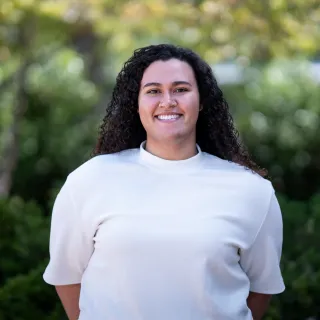Photo of Jayden Sheridan smiling to camera. She has long, curly black hair and is wearing a white short sleeve shirt